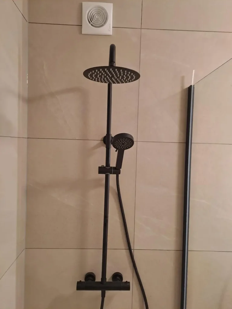 Colonne de douche noire avec pommeau de tête rond et douchette sur un mur carrelé beige.