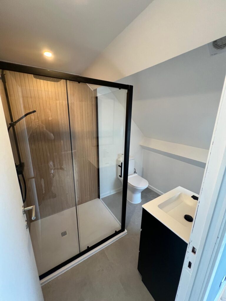 Salle d’eau sous plafond en pente avec douche vitrée, meuble vasque noir et WC.