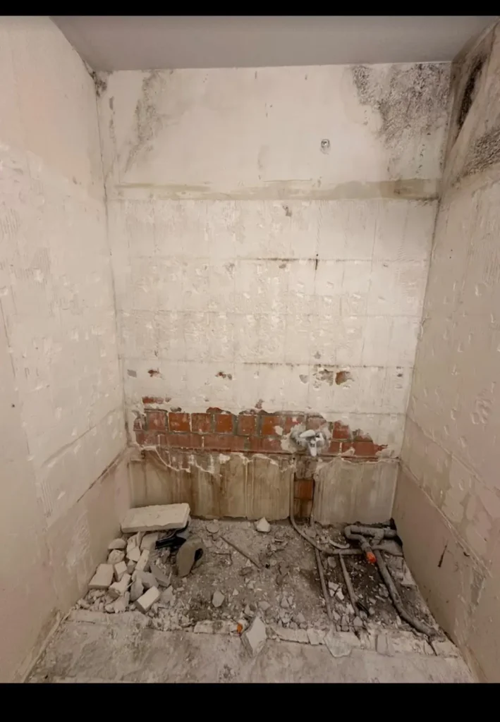 Salle de bain en cours de dépose, avec murs dégarnis, canalisations apparentes et gravats au sol.