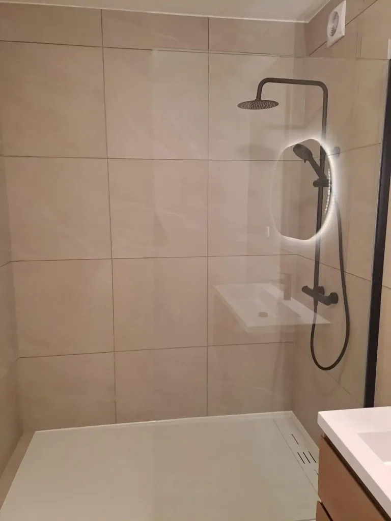 Douche avec receveur blanc, paroi vitrée, colonne noire et carrelage mural beige.