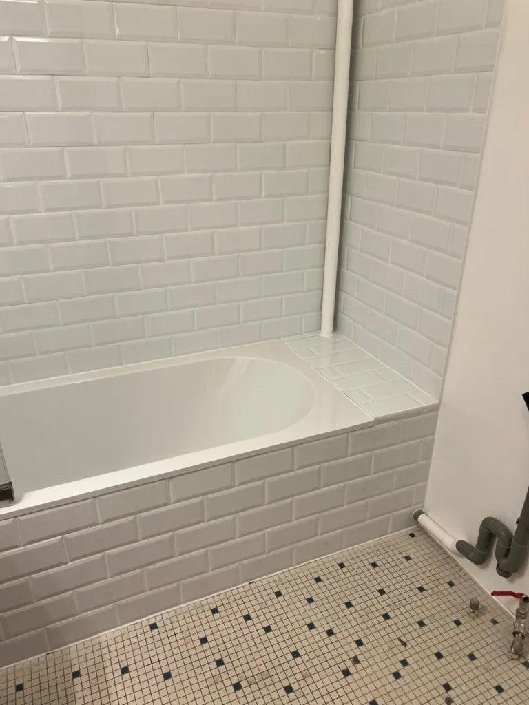 Sol mosaïque clair à motifs foncés au pied d’une baignoire habillée de carrelage blanc.