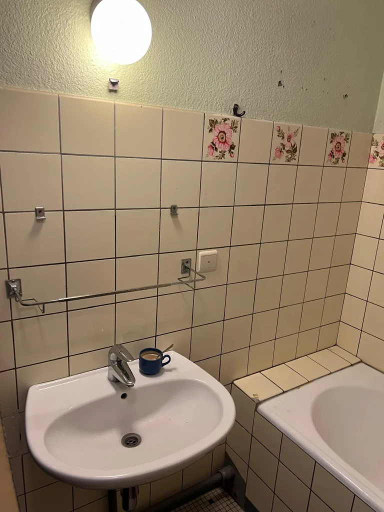 Lavabo mural blanc à côté de la baignoire, devant un carrelage crème à frise florale.