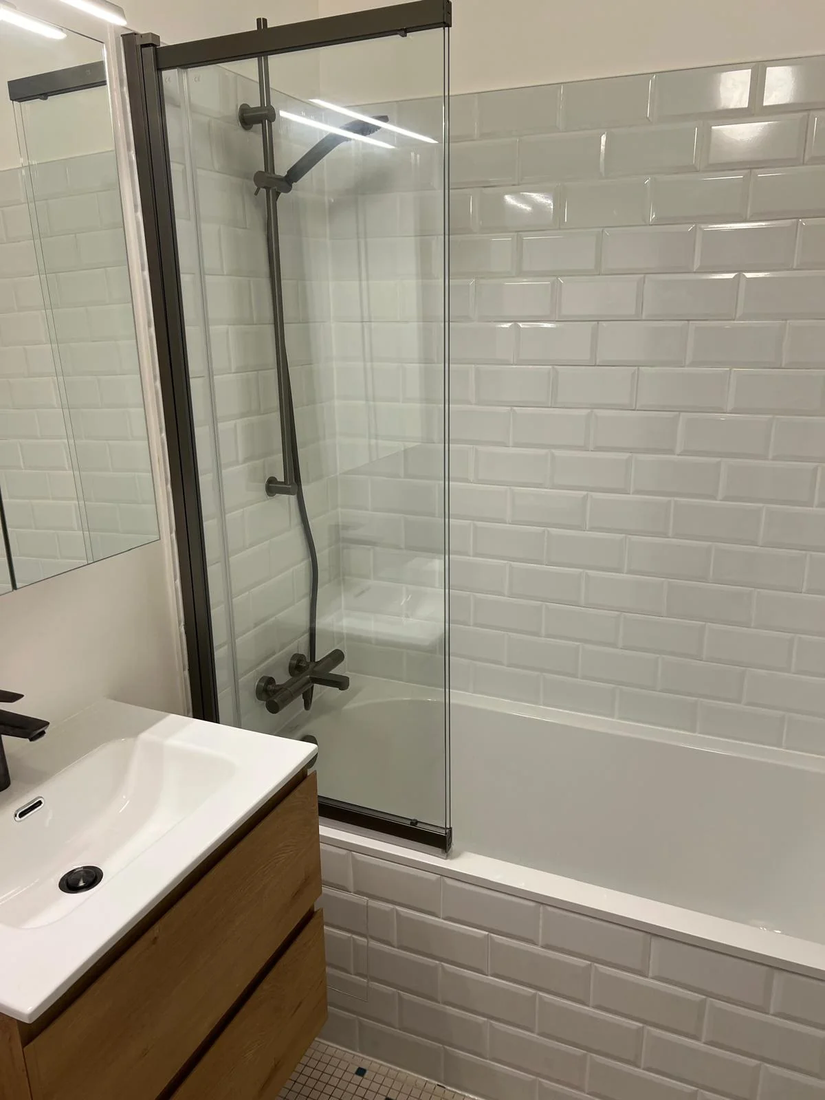 Salle de bain rénovée à Jette Salle de bain rénovée à Jette avec baignoire vitrée, carrelage métro blanc et meuble vasque finition bois.