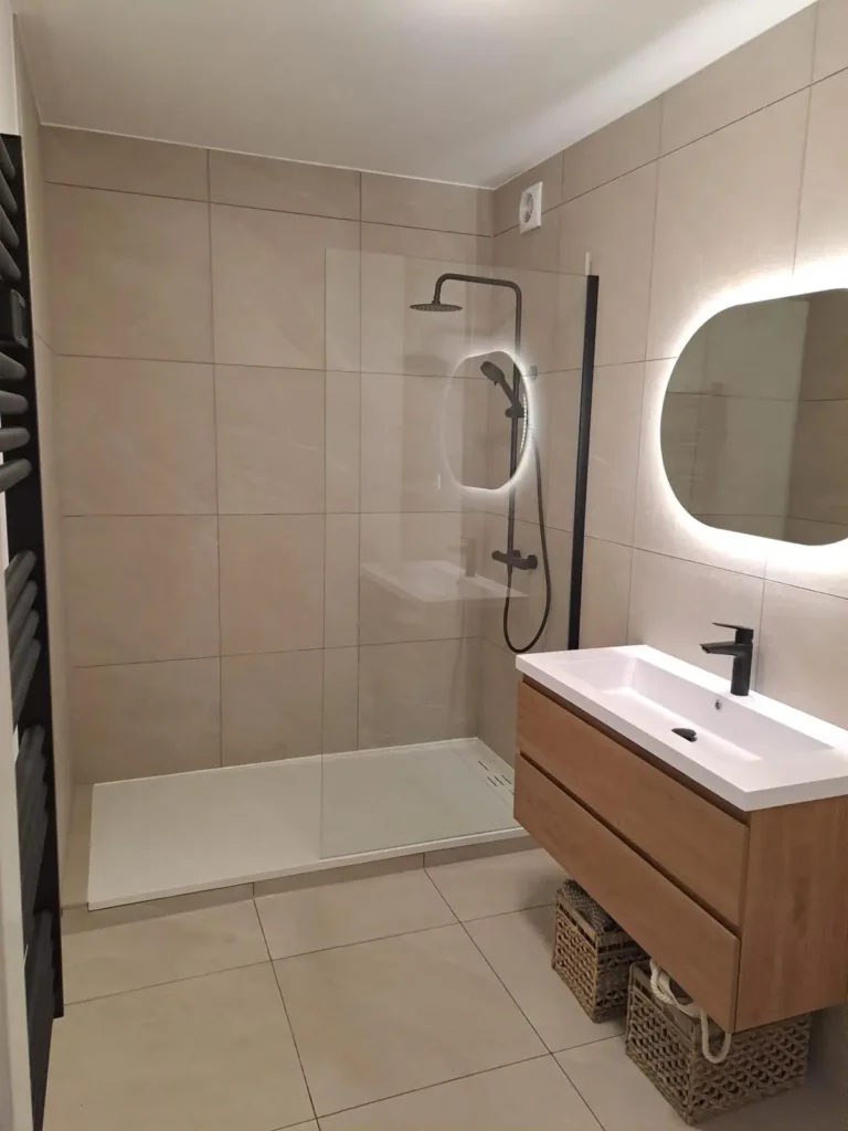 Salle de bain rénovée à Schaerbeek avec douche vitrée, carrelage beige et meuble vasque suspendu finition bois.