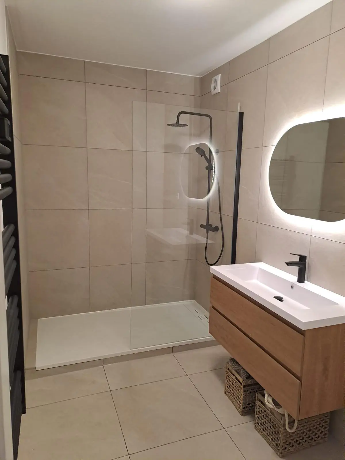 Salle de bain rénovée à Schaerbeek avec douche vitrée, carrelage beige et meuble vasque suspendu finition bois.
