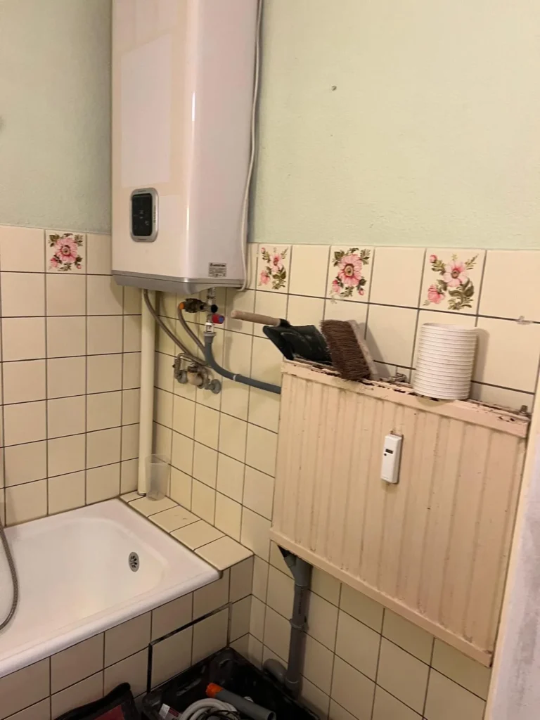 Ancienne salle de bain avec chauffe-eau mural, radiateur et carrelage à motifs floraux.
