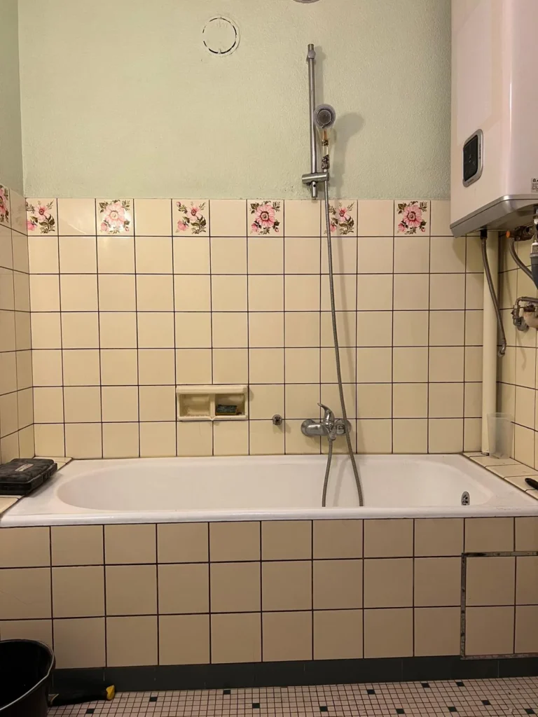 Ancienne baignoire avec carrelage mural crème à motifs floraux et douchette chromée.